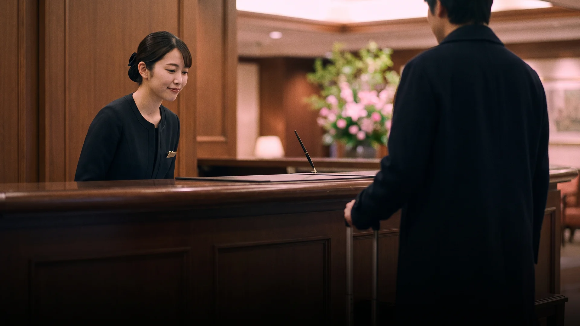 Hotel receptionist in a dark uniform bowing slightly while greeting a guest at a wooden front desk