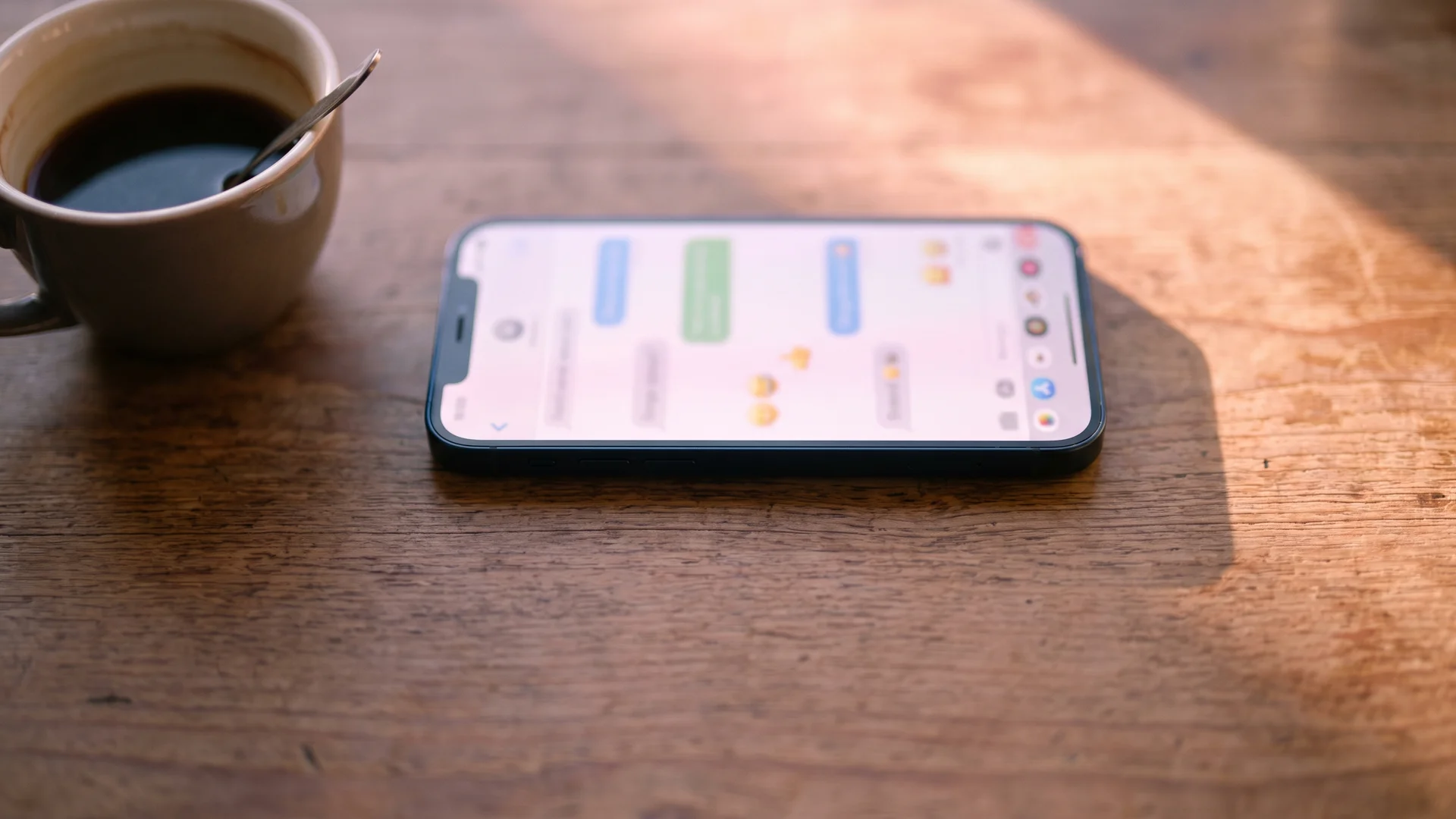 A smartphone lying on a wooden table showing a blurred messaging app chat, with a cup of coffee nearby