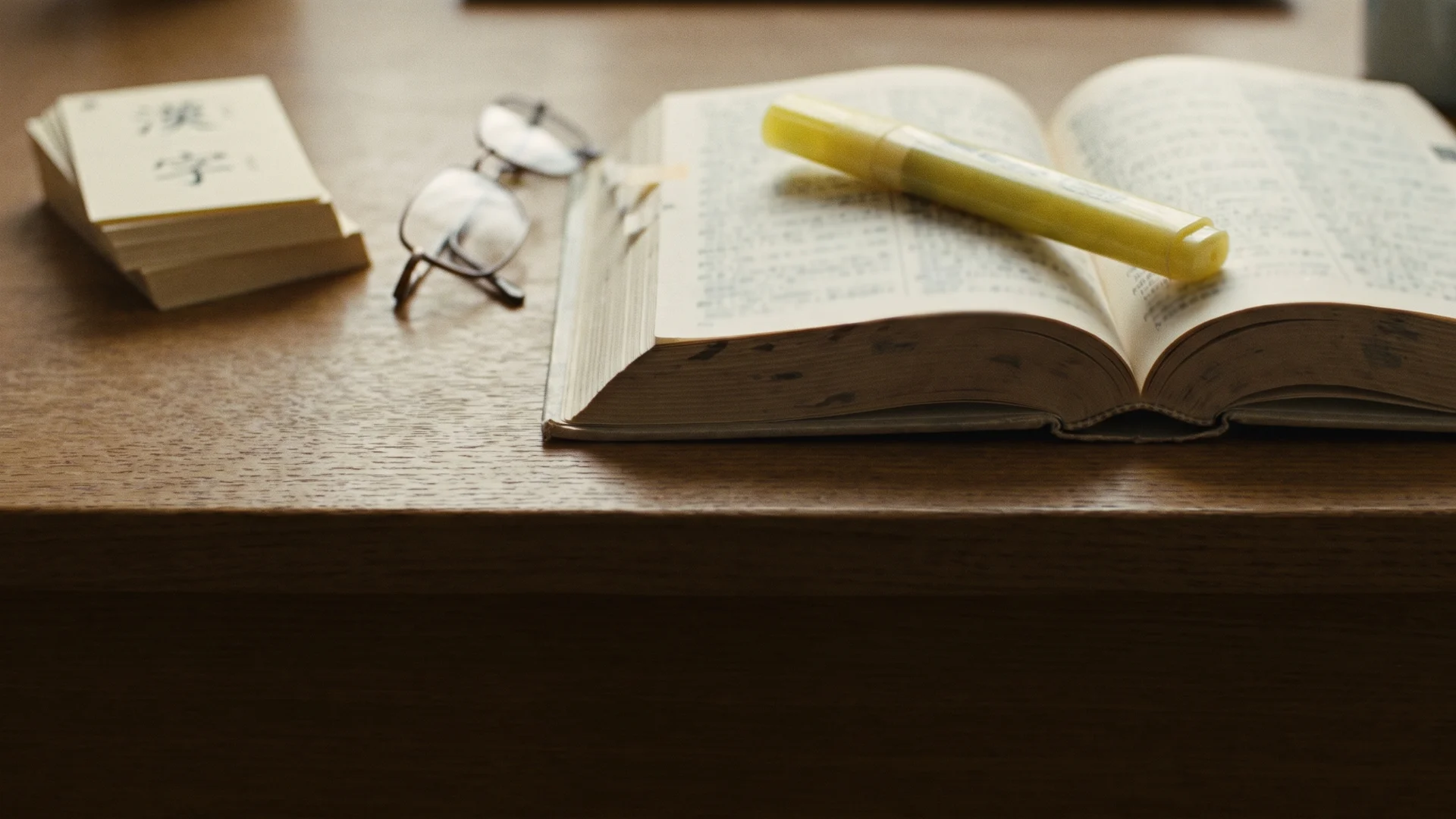 A Japanese dictionary open on a wooden desk with a yellow highlighter resting on the page, a small kanji flashcard stack and reading glasses nearby, no hands in frame