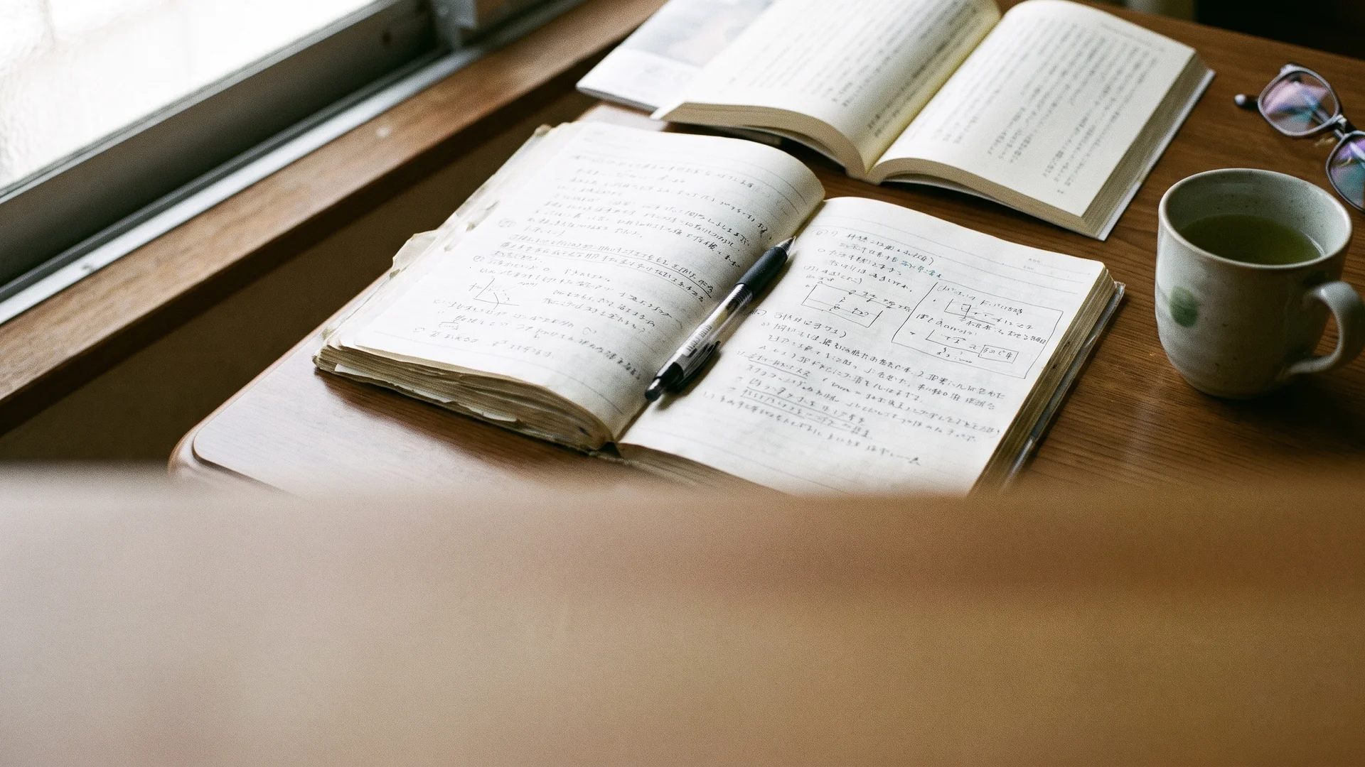 An open notebook with handwritten Japanese grammar notes and a paperback book on a wooden desk, with tea and reading glasses nearby